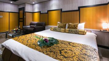 A hotel room with a large bed, a leather armchair, a bedside table with a lamp, and a small table with a fruit bowl and bottle.
