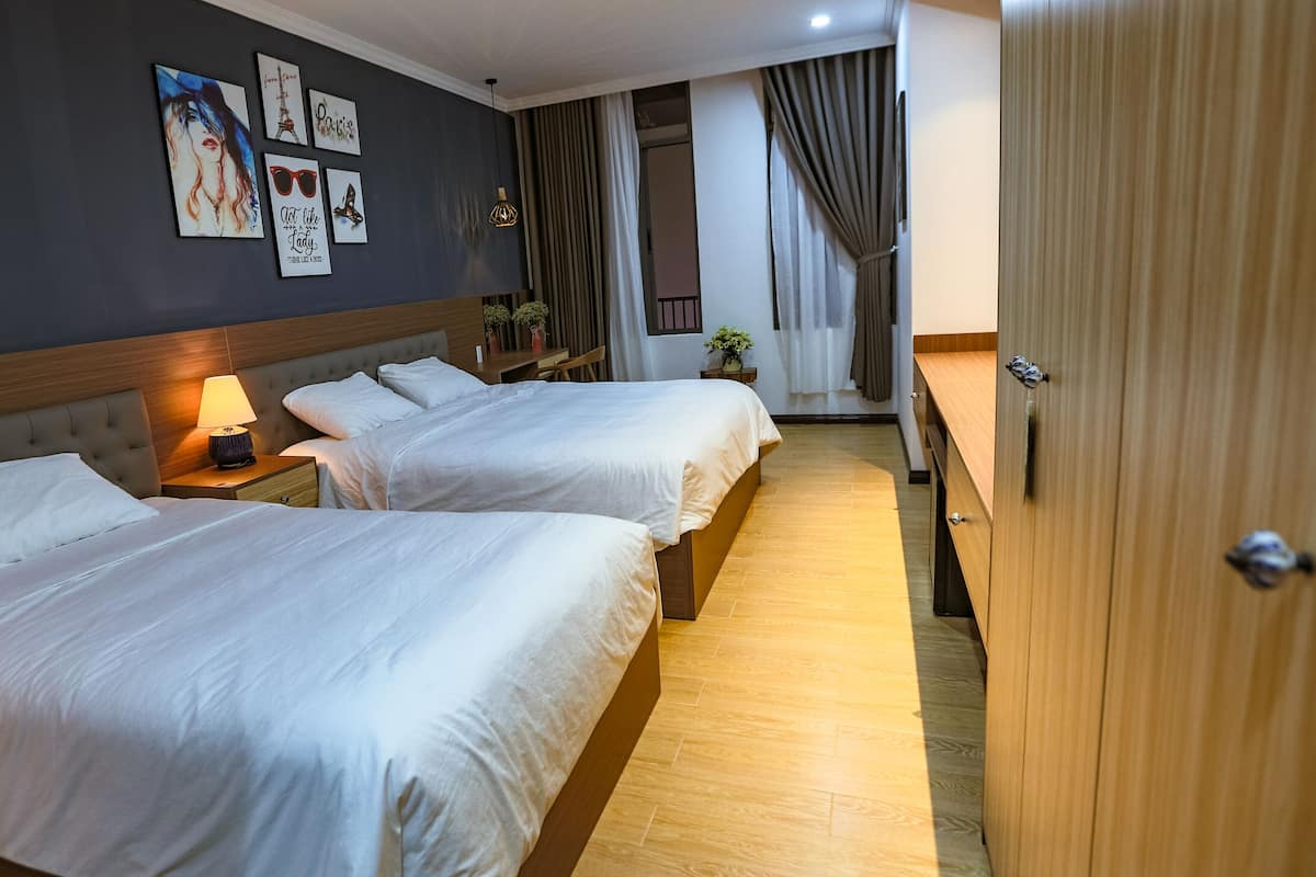 Habitación de hotel con dos camas, una mesita de noche con lámpara, cabecera de madera y una pared decorada con obras de arte.