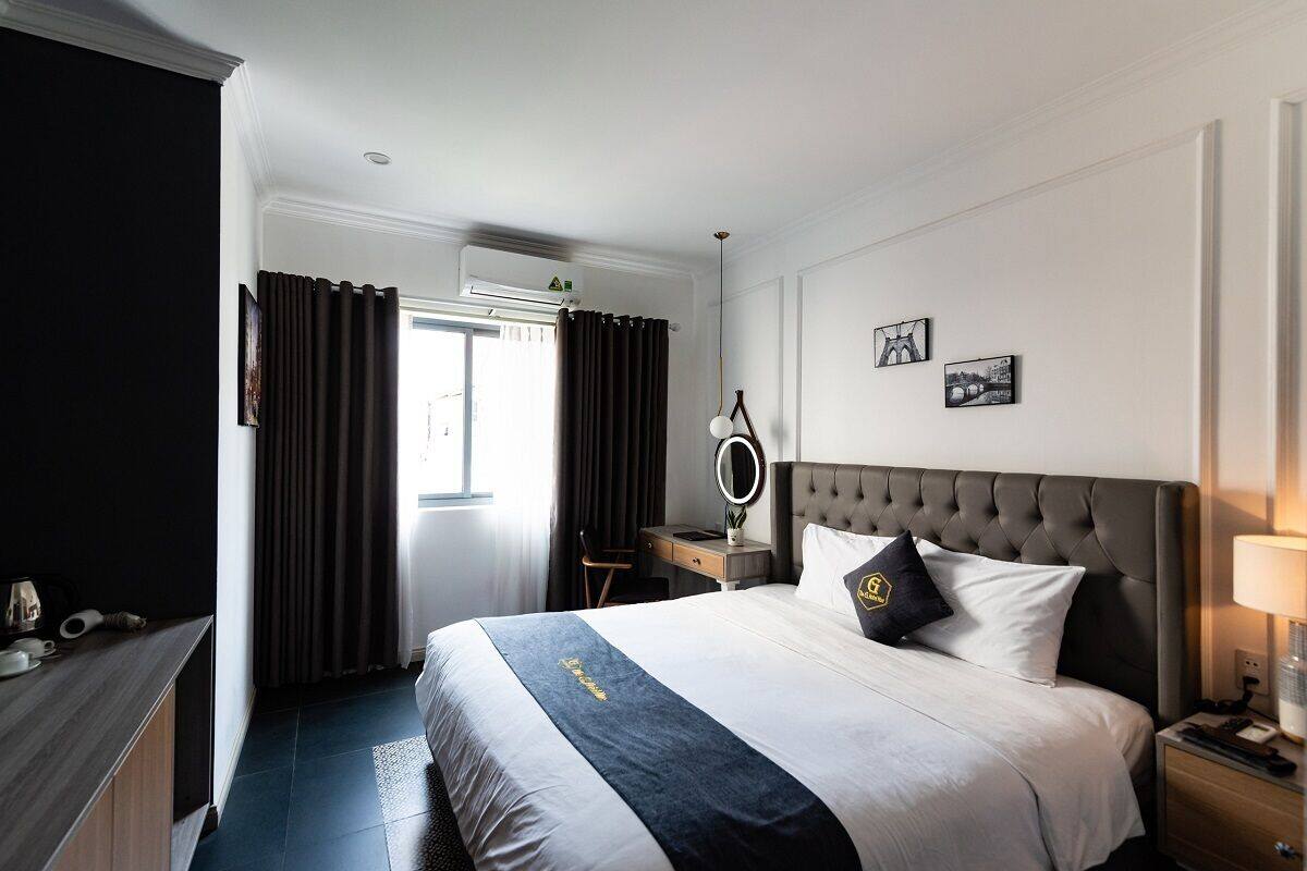 Habitación de hotel con una cama grande, una mesita de noche con lámpara, un escritorio con silla y una ventana con cortinas.