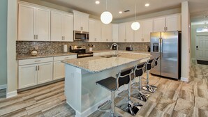 A modern kitchen with white cabinets, a granite countertop, stainless steel appliances, and a bar area with stools. - Champions Gate Home 2 U- Maui- Cg1560 (Davenport)