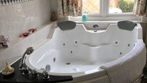A bathroom with a large bathtub, a toilet, and a window with curtains. - West End Lodge (Esher)