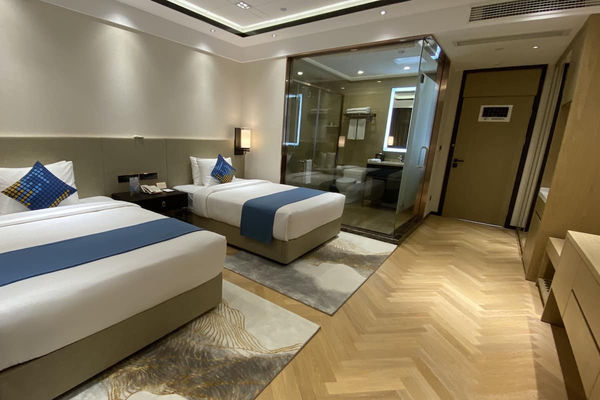 Habitación de hotel con dos camas, un espejo grande, un baño visible a través de una puerta de vidrio y suelo de madera.