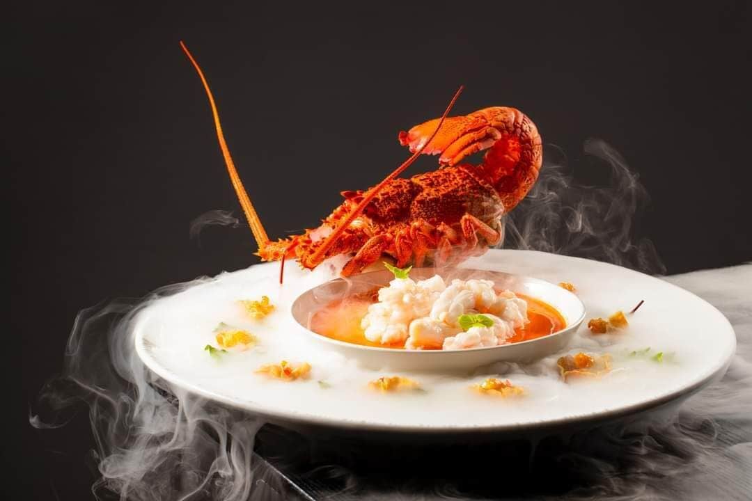 A lobster on a plate with a bowl of soup and some garnish.