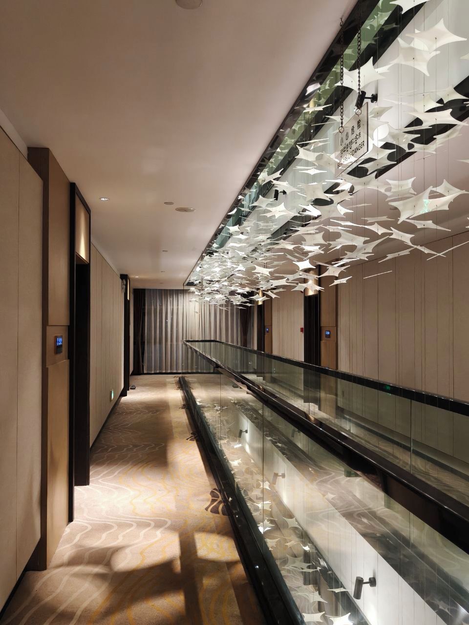 A modern hotel corridor with a glass railing, a patterned carpet, and a ceiling with hanging light fixtures.