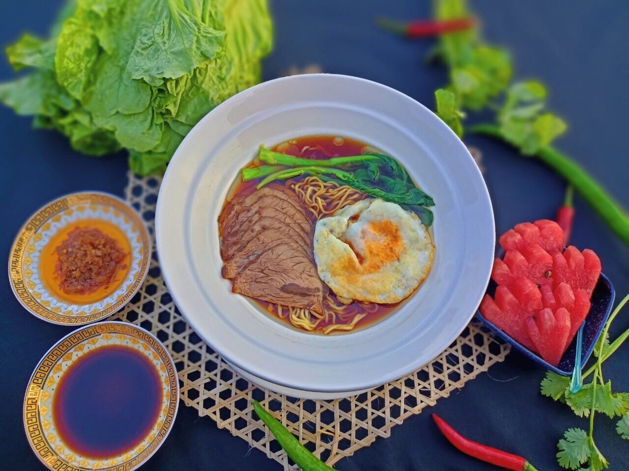 A bowl of noodle soup with beef and egg, surrounded by vegetables and condiments.