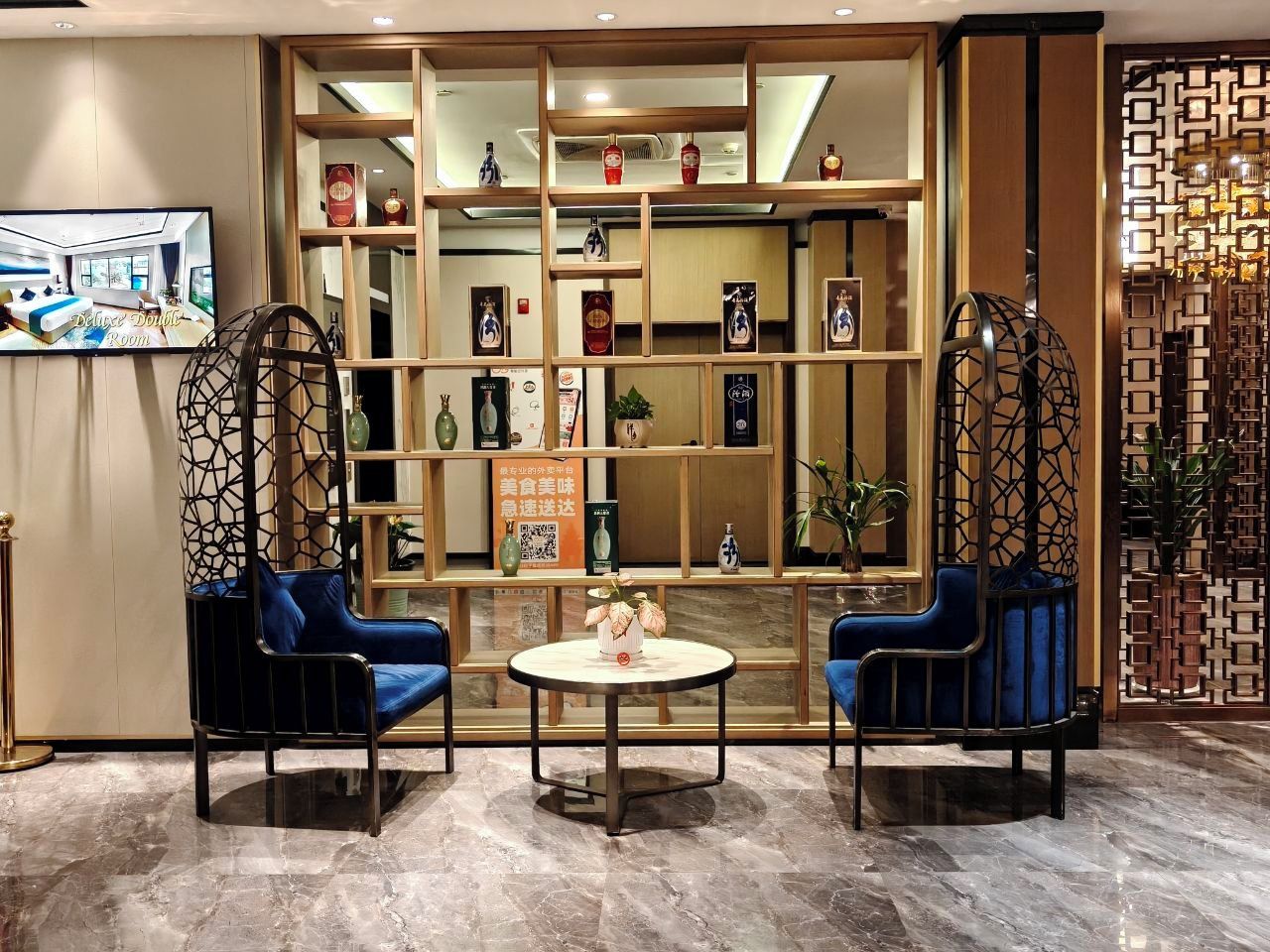 A hotel lobby with a reception desk, a seating area with two blue chairs, a round table, and a decorative wall with shelves.