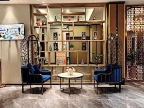 A hotel lobby with a reception desk, a seating area with two blue chairs, a round table, and a decorative wall with shelves.