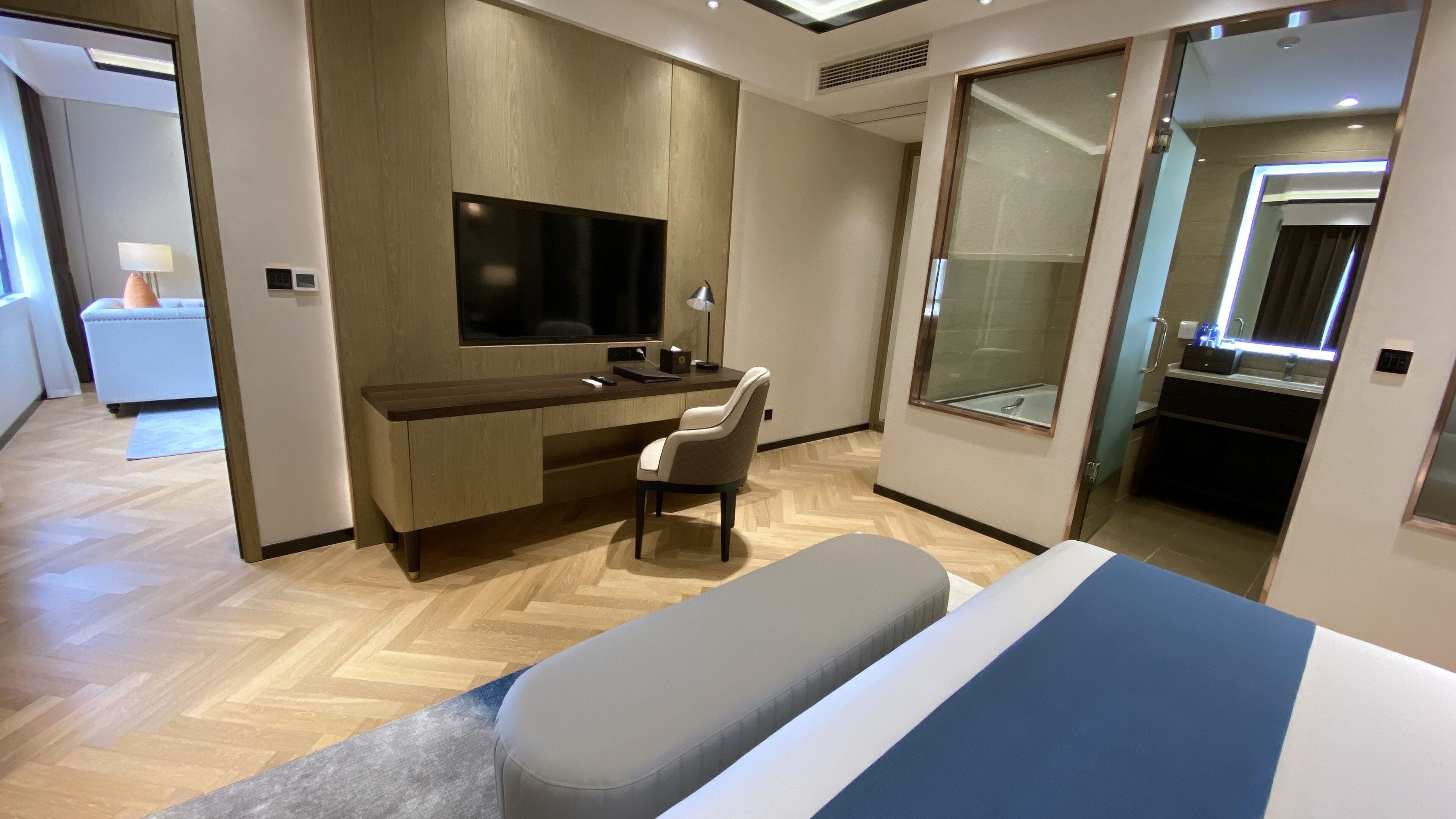Une chambre d’hôtel moderne avec un plancher en bois, un téléviseur à écran plat fixé au mur, un bureau avec une chaise, un grand miroir et un lit recouvert d’une courtepointe à rayures bleues et blanches.