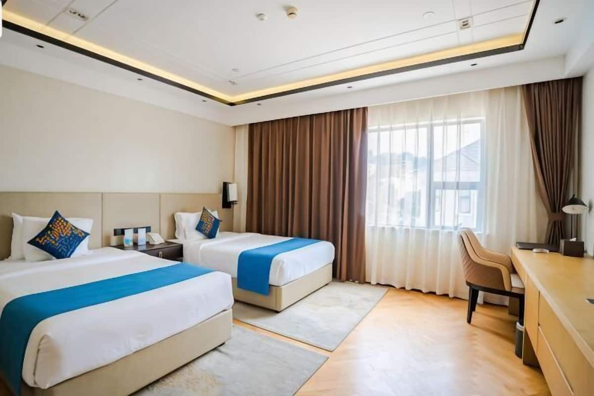Habitación de hotel con dos camas, un escritorio, una silla y un ventanal con cortinas.