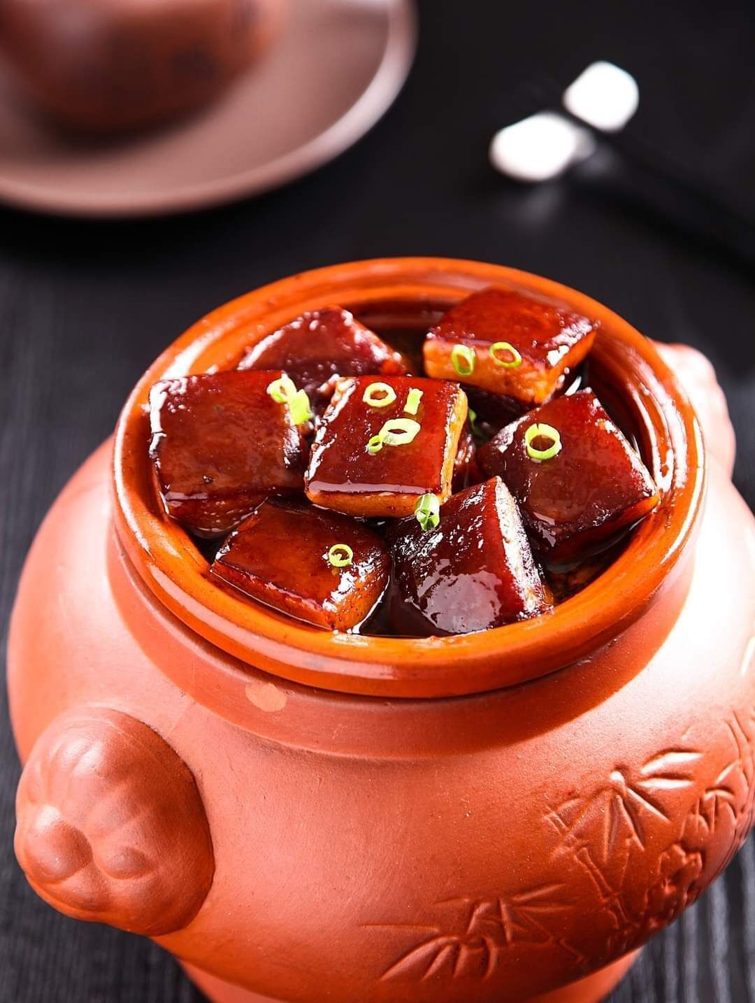 A terracotta pot filled with glazed meat pieces garnished with green onions.