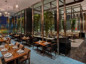 A modern restaurant with large windows, indoor plants, and a patterned floor. - Mövenpick Diyarbakir (Diyarbakir)