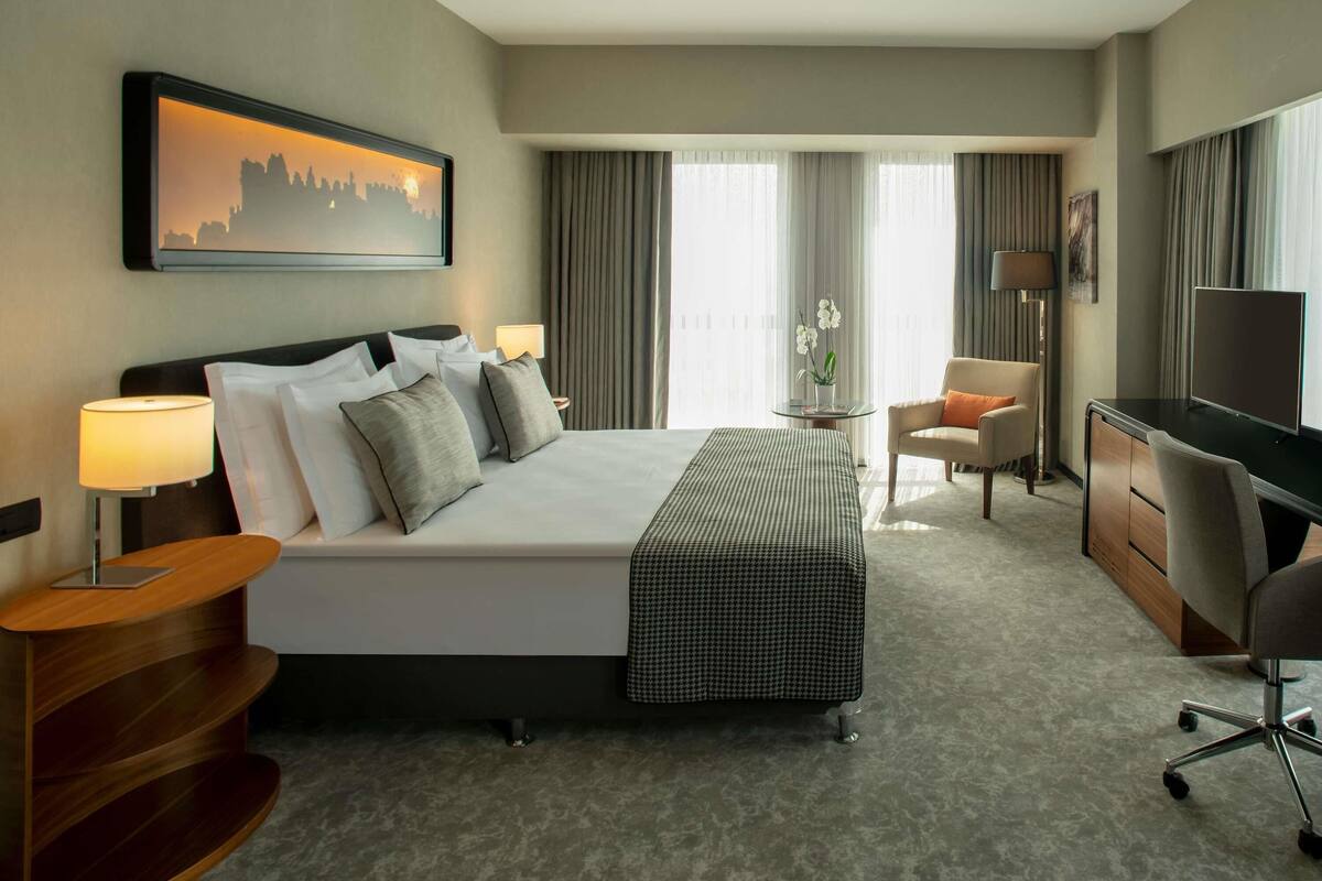 A hotel room with a bed, desk, chair, TV, lamp, and a painting on the wall.