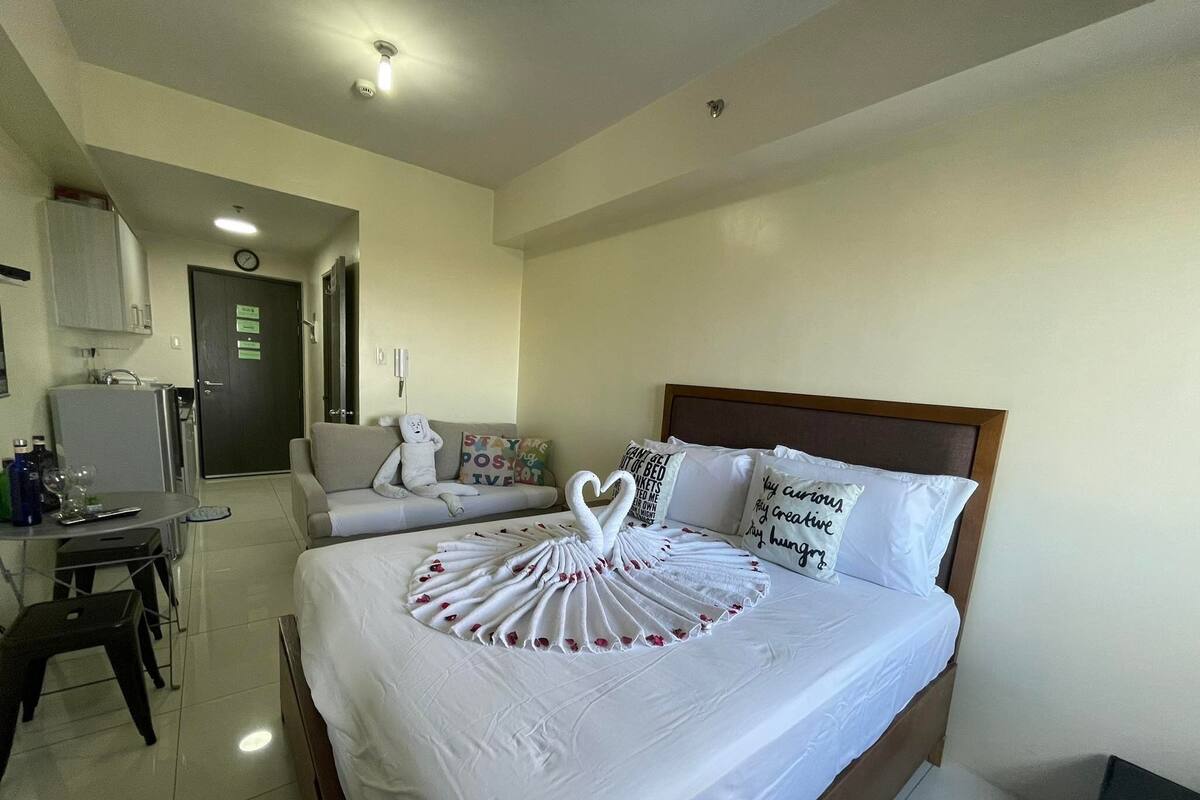 Una habitación de hotel con una cama bien hecha, un escritorio pequeño con una silla, un refrigerador y una puerta con un cartel.