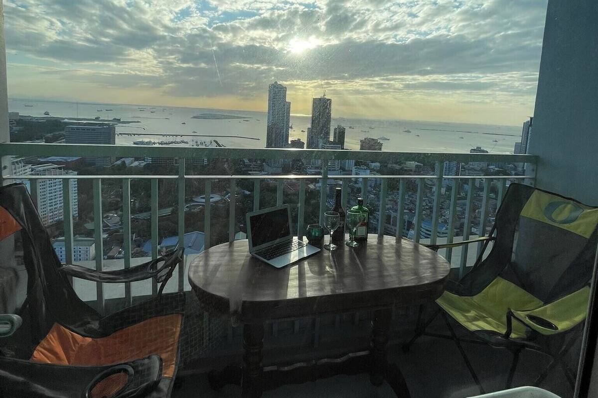 Un balcón con vistas al horizonte urbano y al mar. Hay una mesa con una computadora portátil, una botella y un vaso. Dos sillas están colocadas alrededor de la mesa.