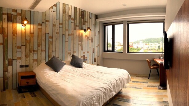 A modern bedroom with a large bed, a desk, and a view of the city through large windows. - The Upstairs (Hualien City)
