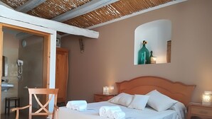 A bedroom with a bed, a chair, a nightstand, a mirror, and a bathroom visible through an open door. - TerraMater (Noto)