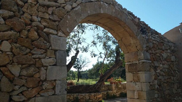 An ancient stone archway leading to a garden with trees and potted plants. - TerraMater (Noto)