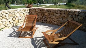 Two wooden lounge chairs on a gravel surface with a stone wall backdrop and a view of a green landscape. - TerraMater (Noto)