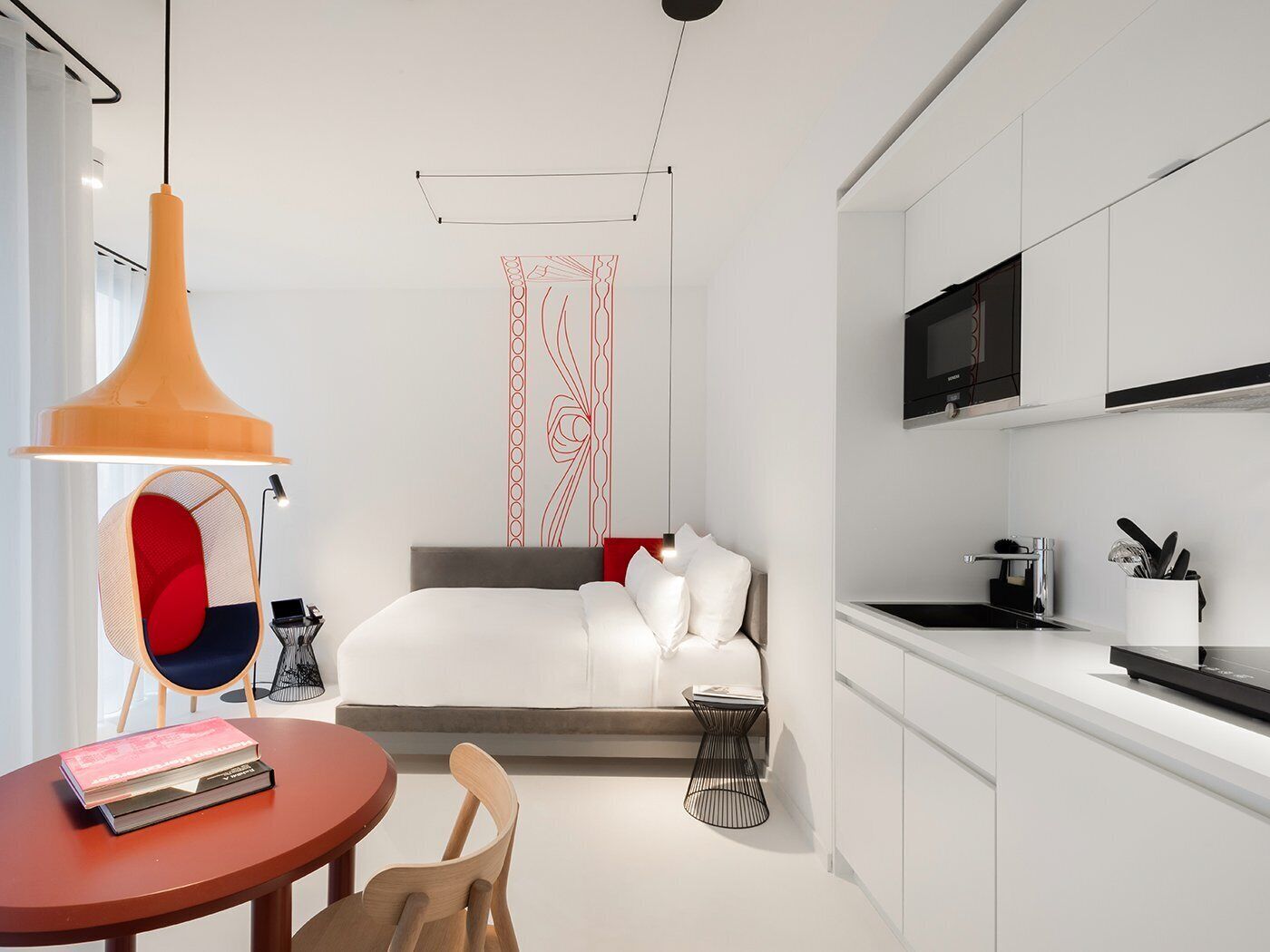 A modern hotel room with a bed, a small table with books, a chair, a kitchen area with a TV, and a unique red and white wall decoration.