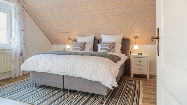 Une chambre avec un plafond en pente, un lit recouvert d’une literie blanche, deux tables de chevet avec des lampes et un tapis à motifs.