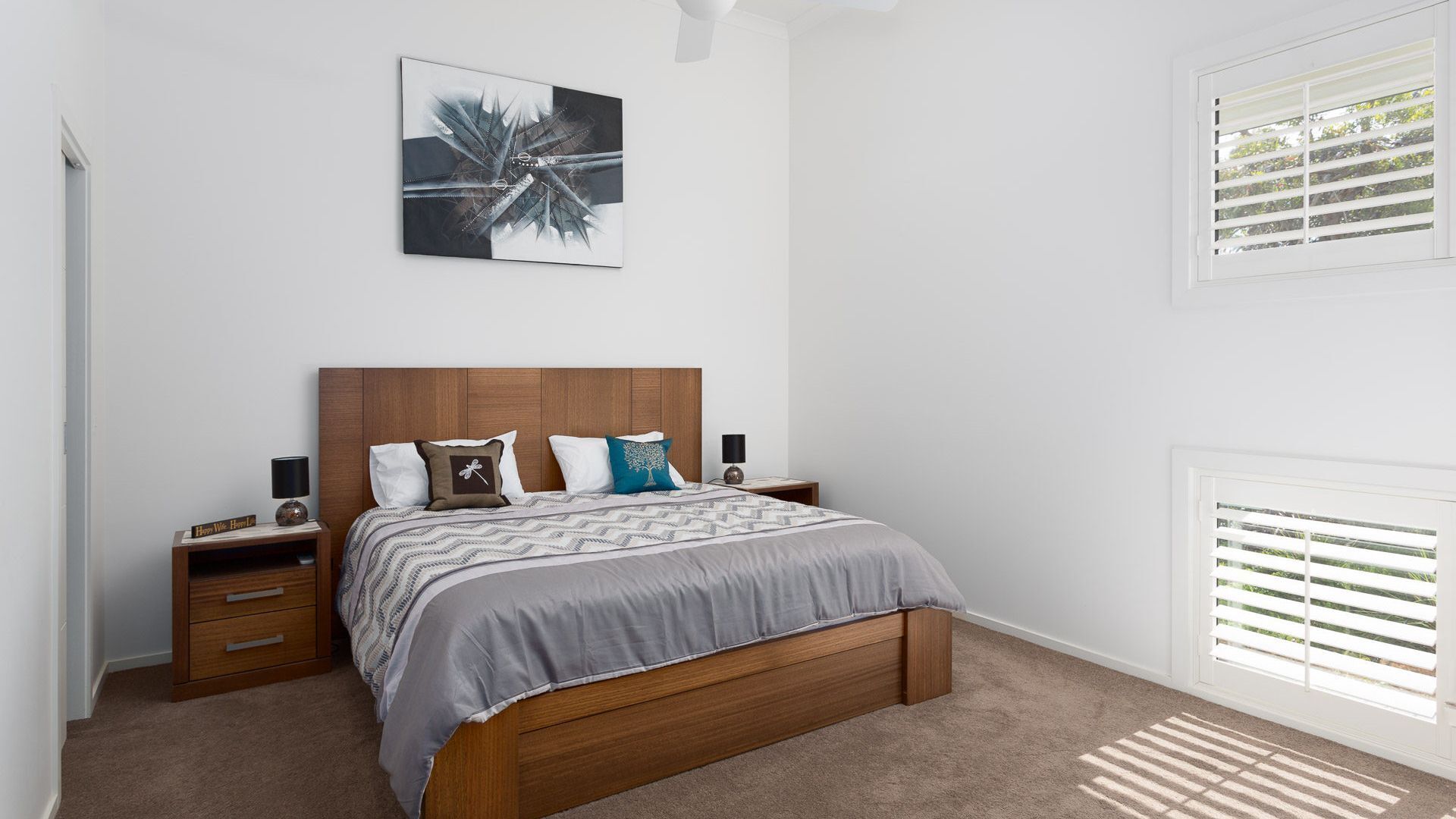 A bedroom with a wooden bed, a nightstand, a bedside table, a wall-mounted artwork, and a window with blinds.