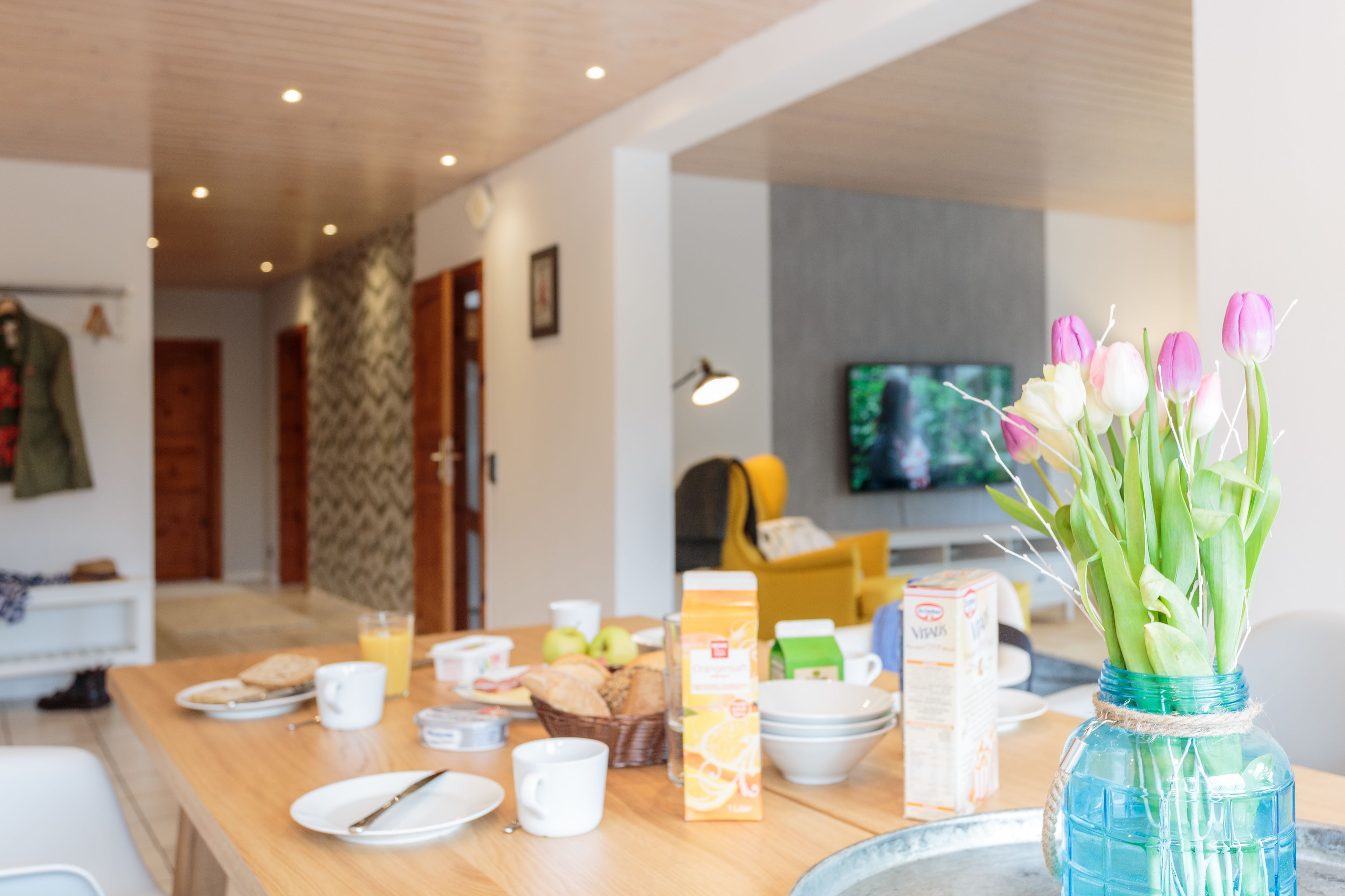 A dining area with a wooden table set for breakfast, including plates, cups, and a vase with flowers.