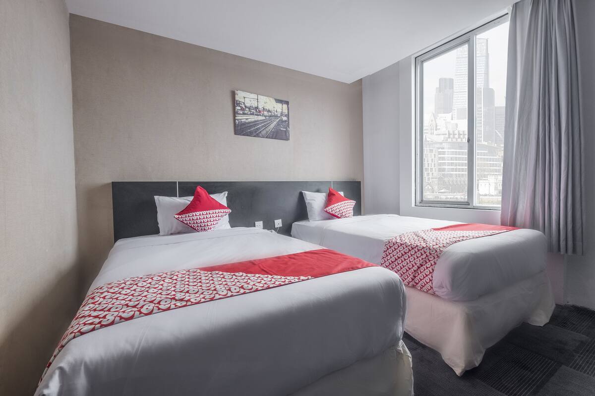 Ein Hotelzimmer mit zwei Betten, jeweils mit roter und weißer Bettwäsche, Blick auf die Stadtlandschaft durch ein großes Fenster und einem gerahmten Bild an der Wand.