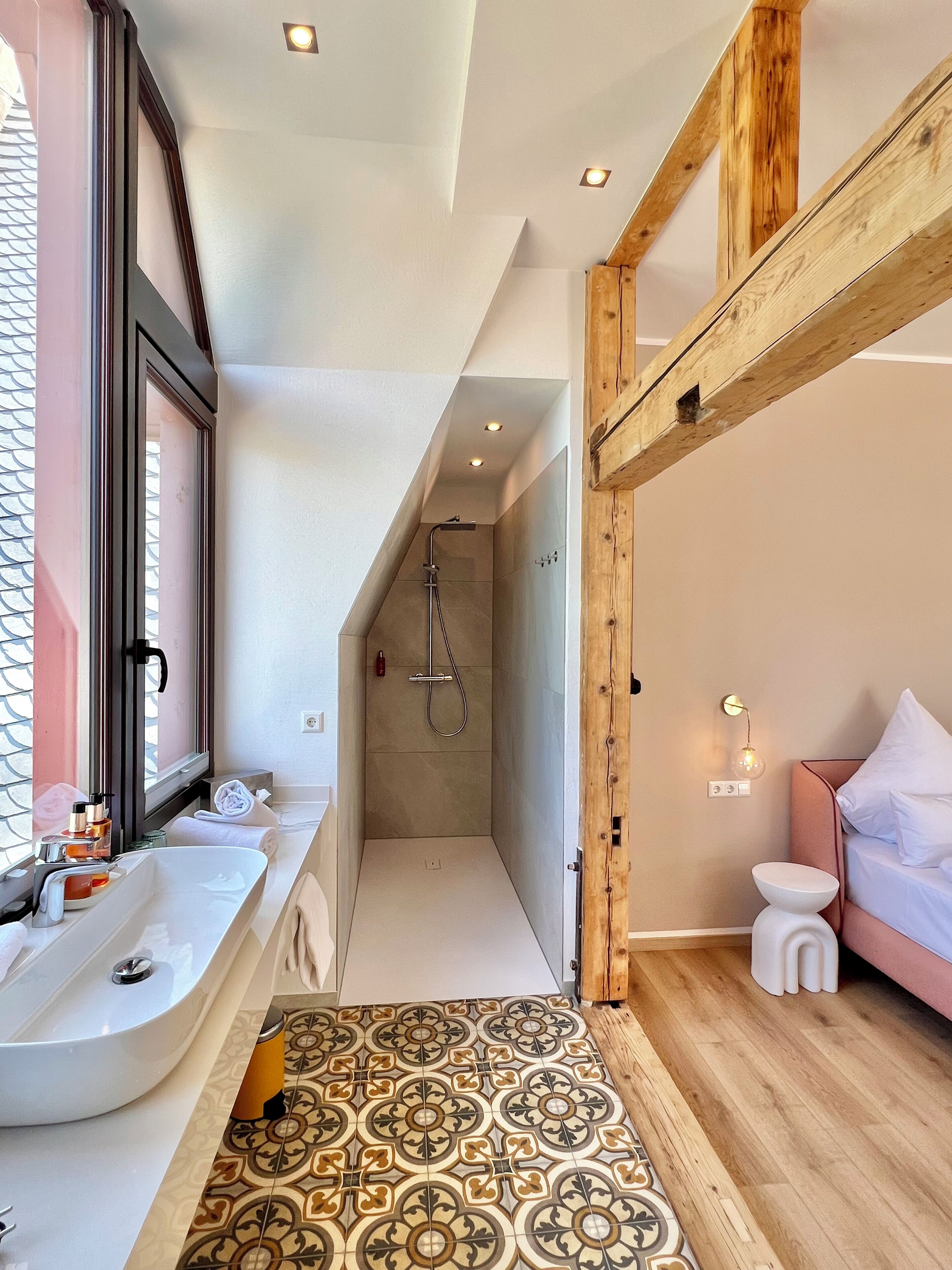 A modern bathroom with a large mirror, two sinks, a shower, and a wooden ceiling beam.