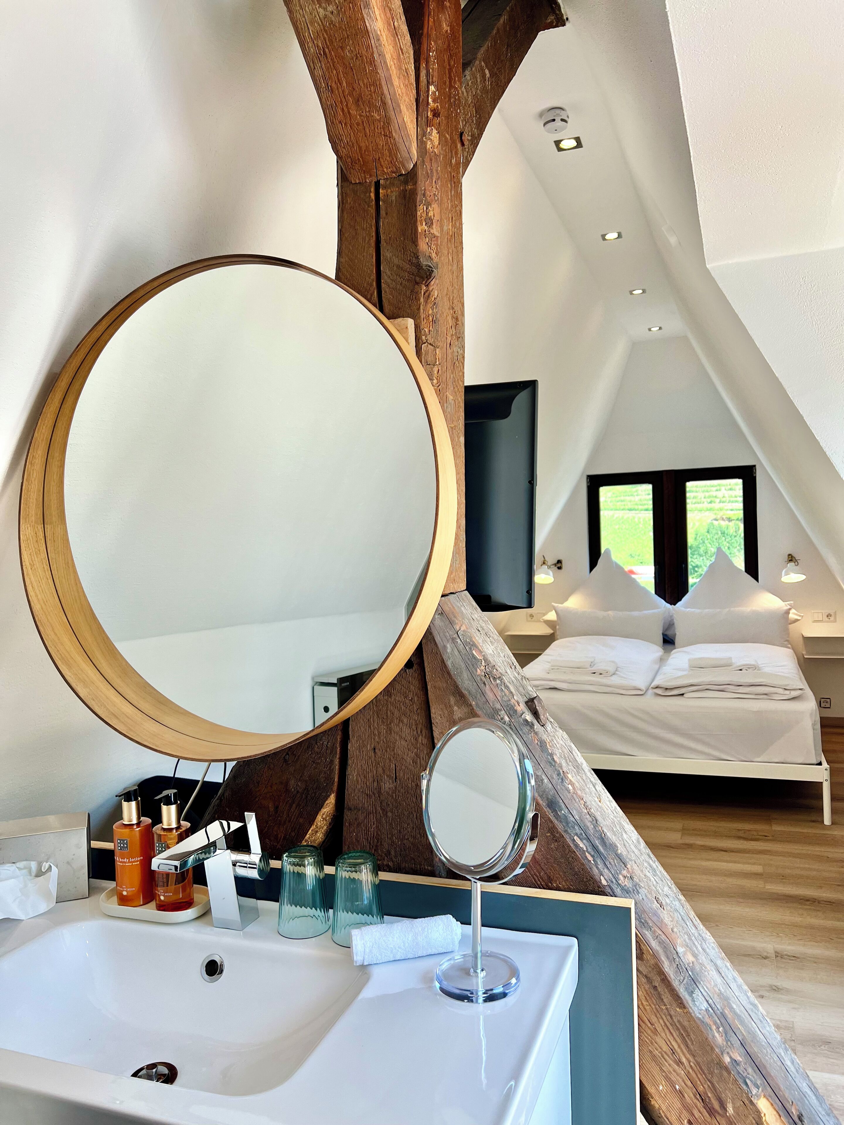 A bedroom with a bed, a large mirror, and a bathroom with a sink and toiletries.