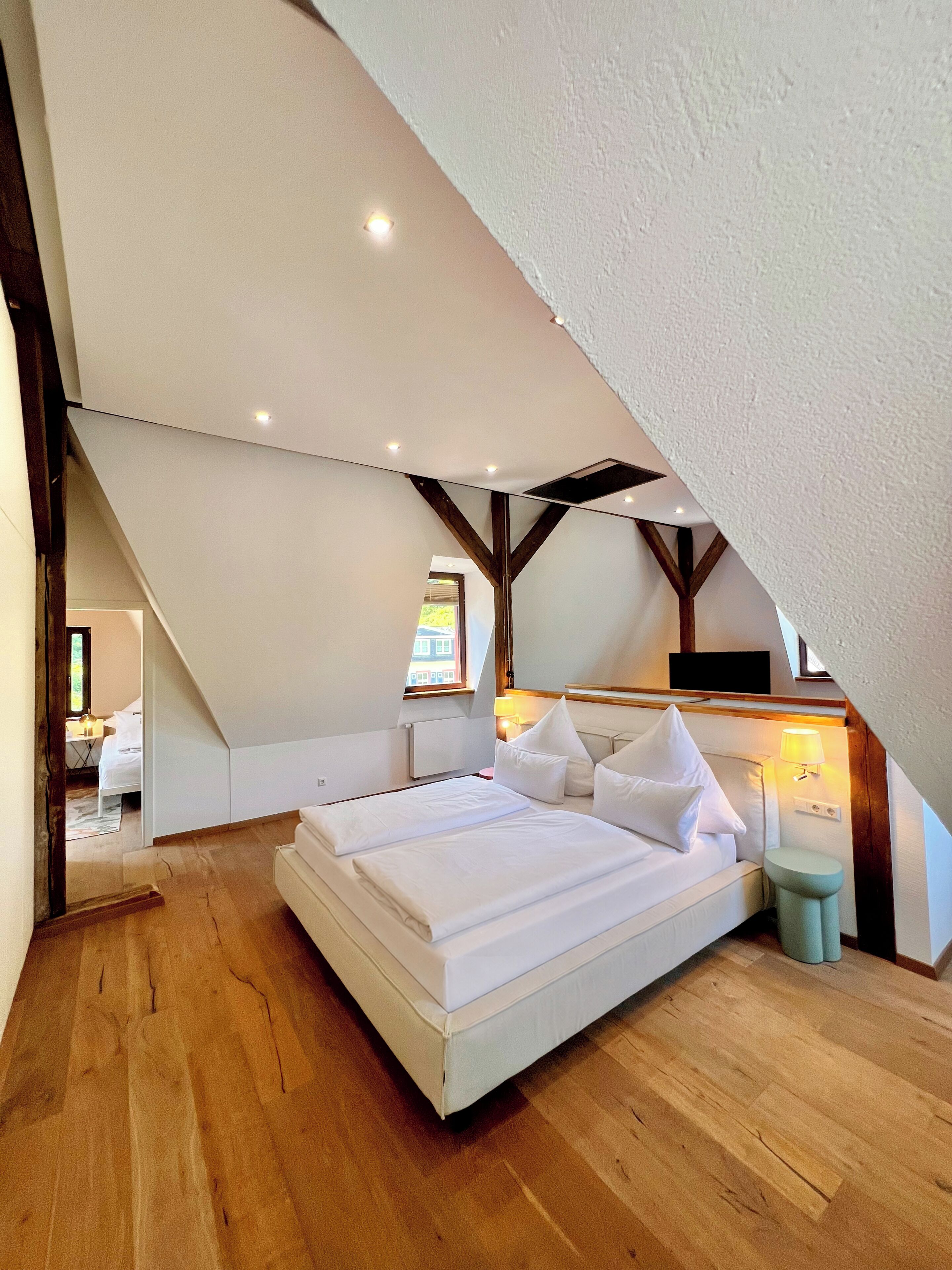 Ein Schlafzimmer mit schrägen Wänden, einem Bett mit weißer Bettwäsche, einem Nachttisch mit Lampe und einem Fenster mit Ausblick.