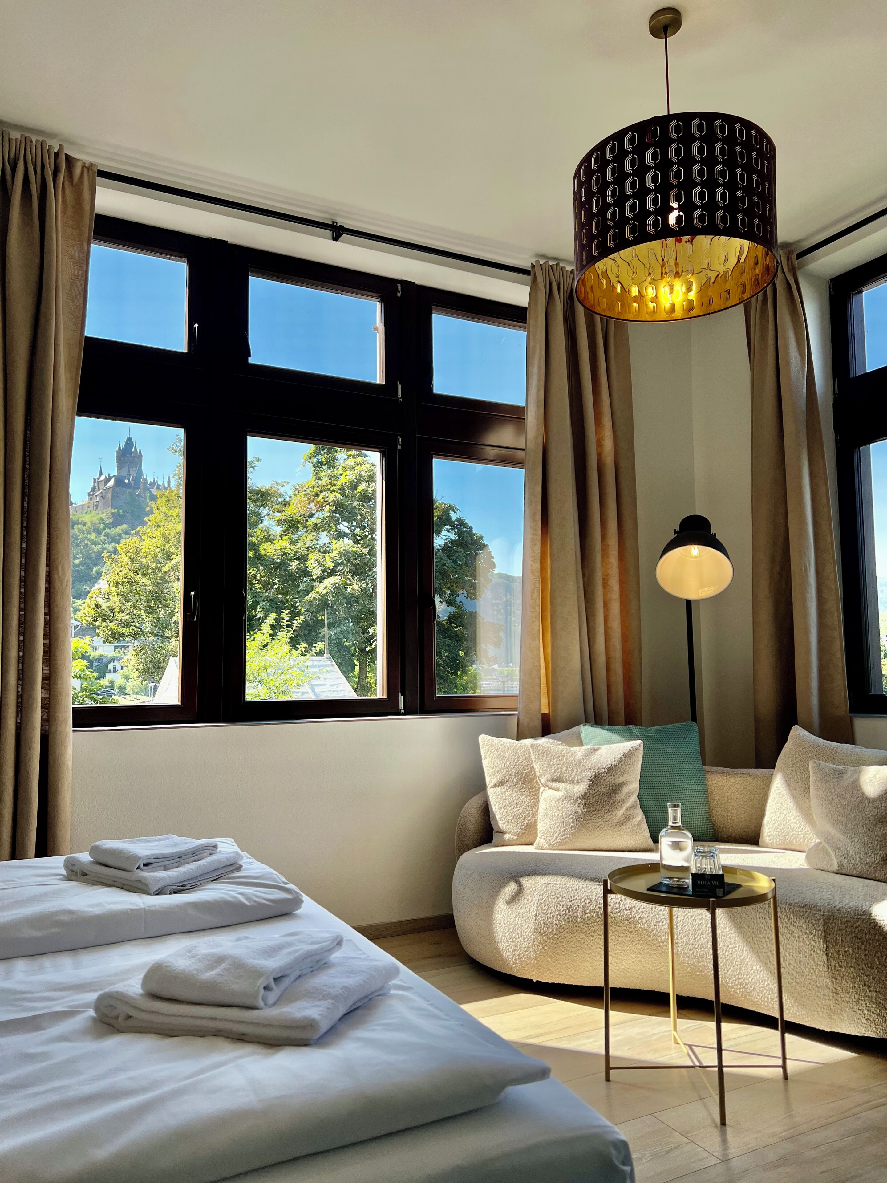 Een hotelkamer met een bed, een sofa, een tafel en grote ramen met uitzicht op een kasteel.