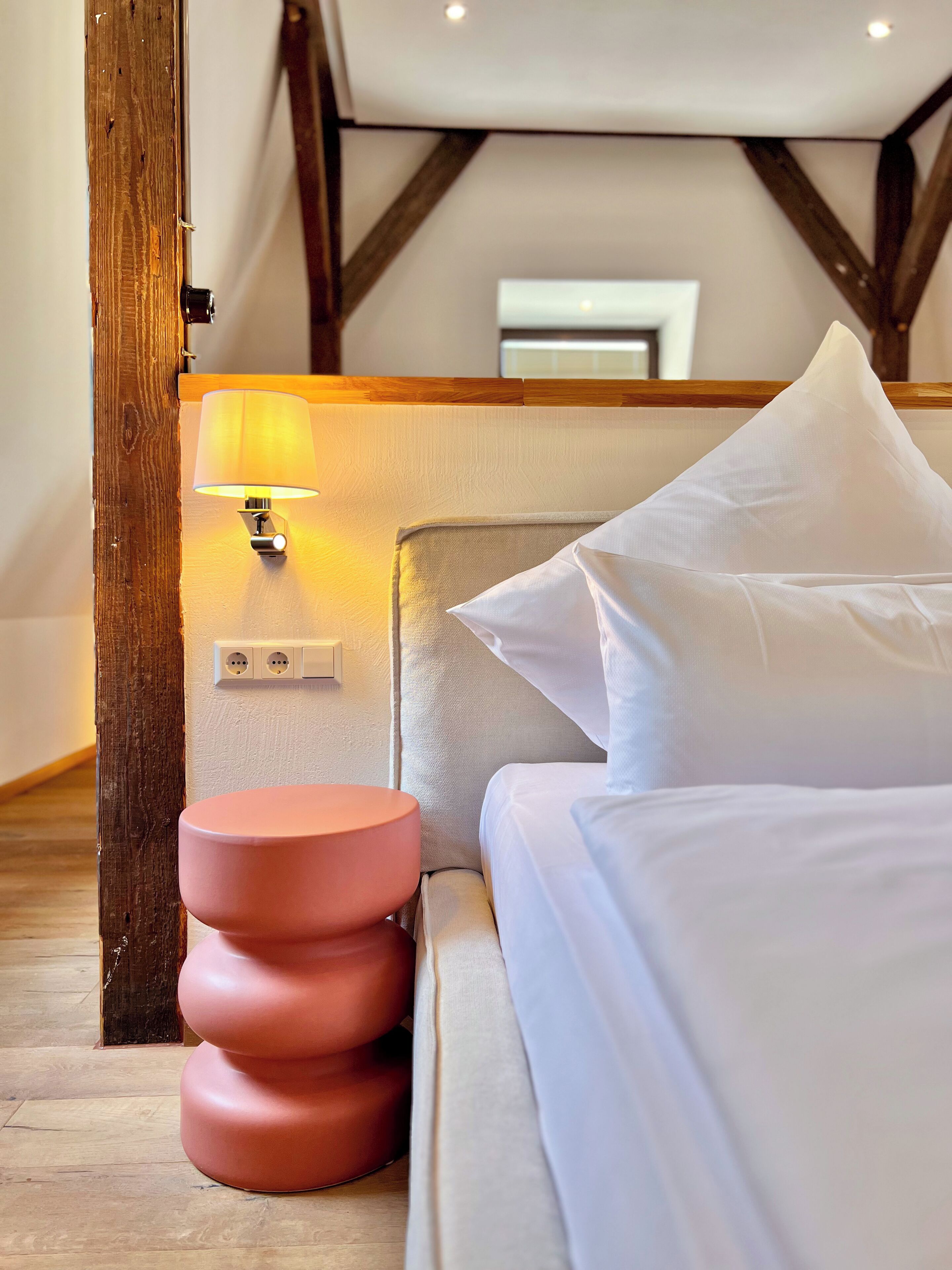 A hotel room with a bed, a bedside lamp, a round pink nightstand, and a wooden beam structure.