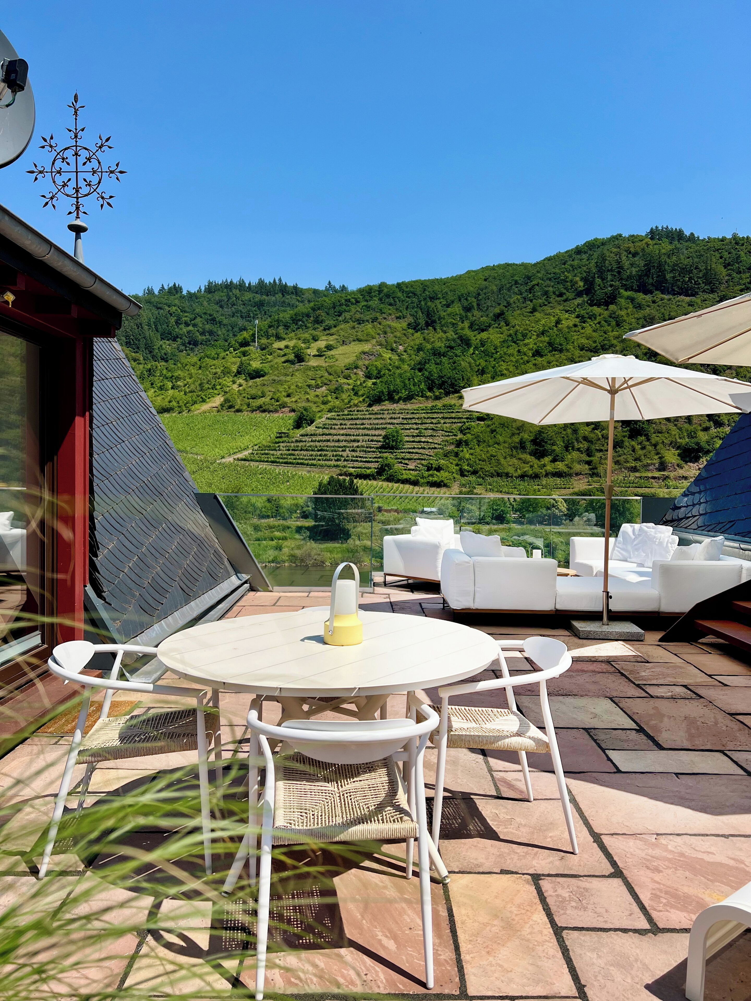 Paseo con mobiliario exterior, una mesa y sombrillas, con vistas a un paisaje verde y colinas.