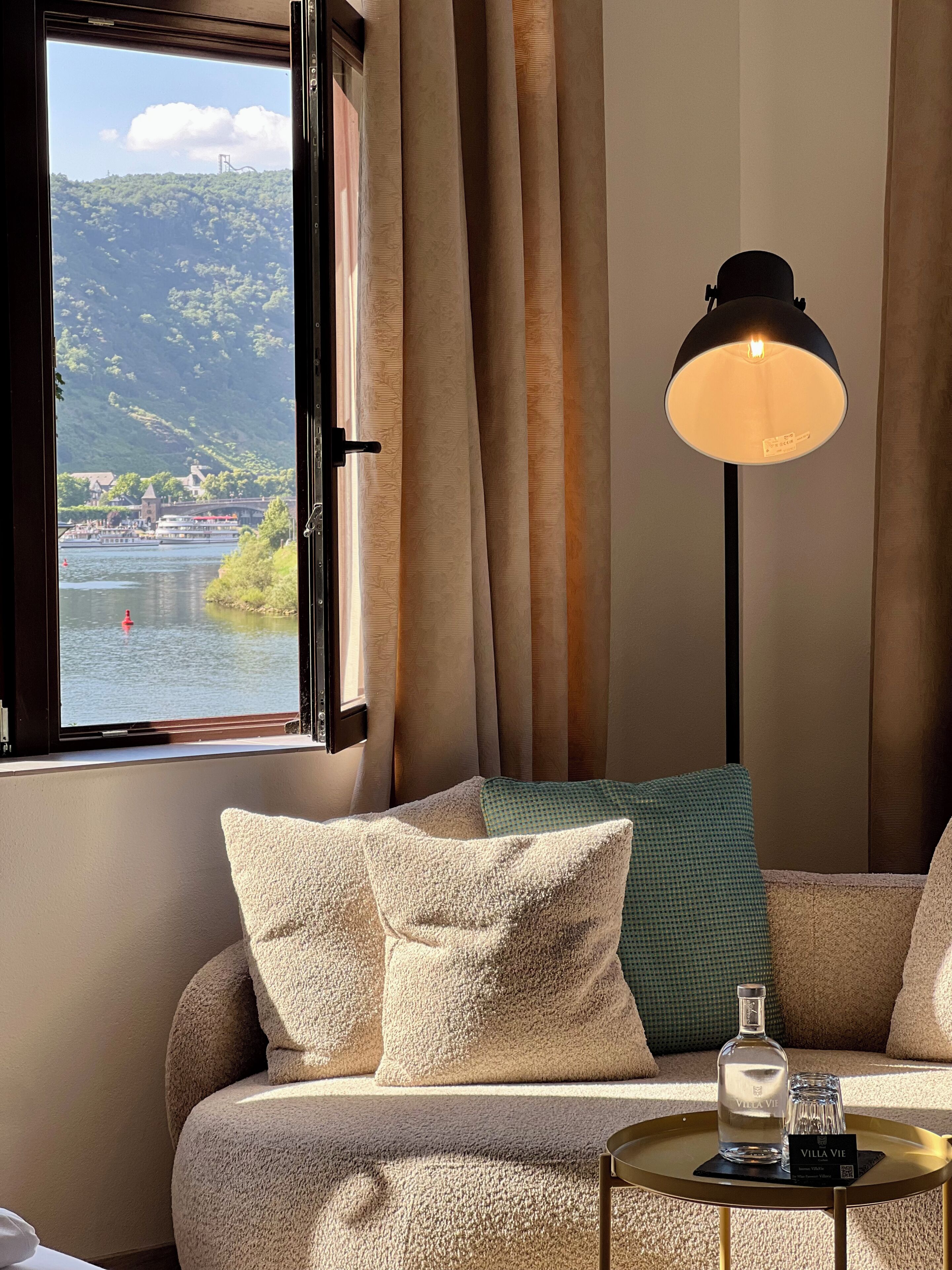 Une pièce avec un canapé, une lampe, une table avec une bouteille et des verres, et une vue sur un lac et des bâtiments.