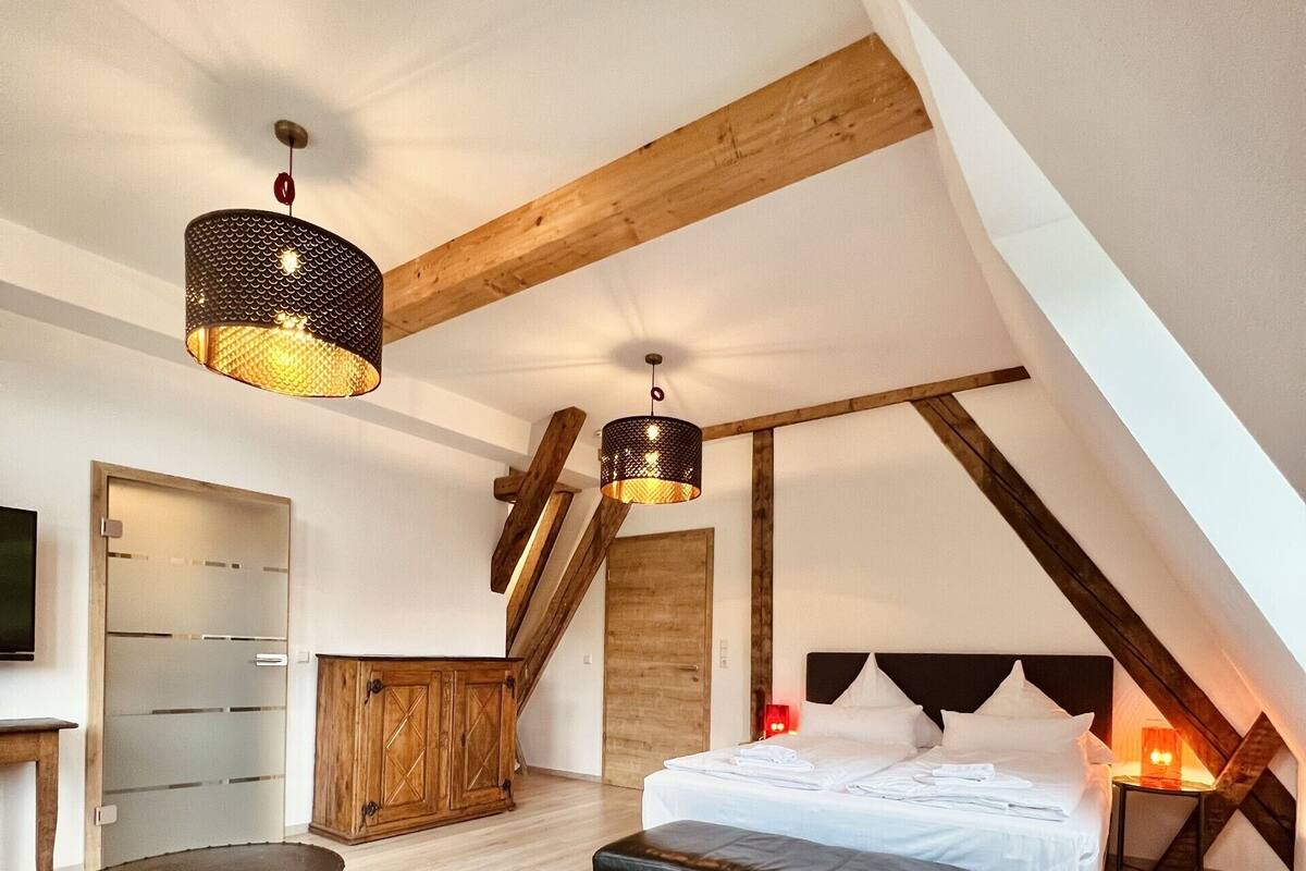 A bedroom with a sloped ceiling, wooden beams, a bed with white linens, a red patterned sofa, and a drum.