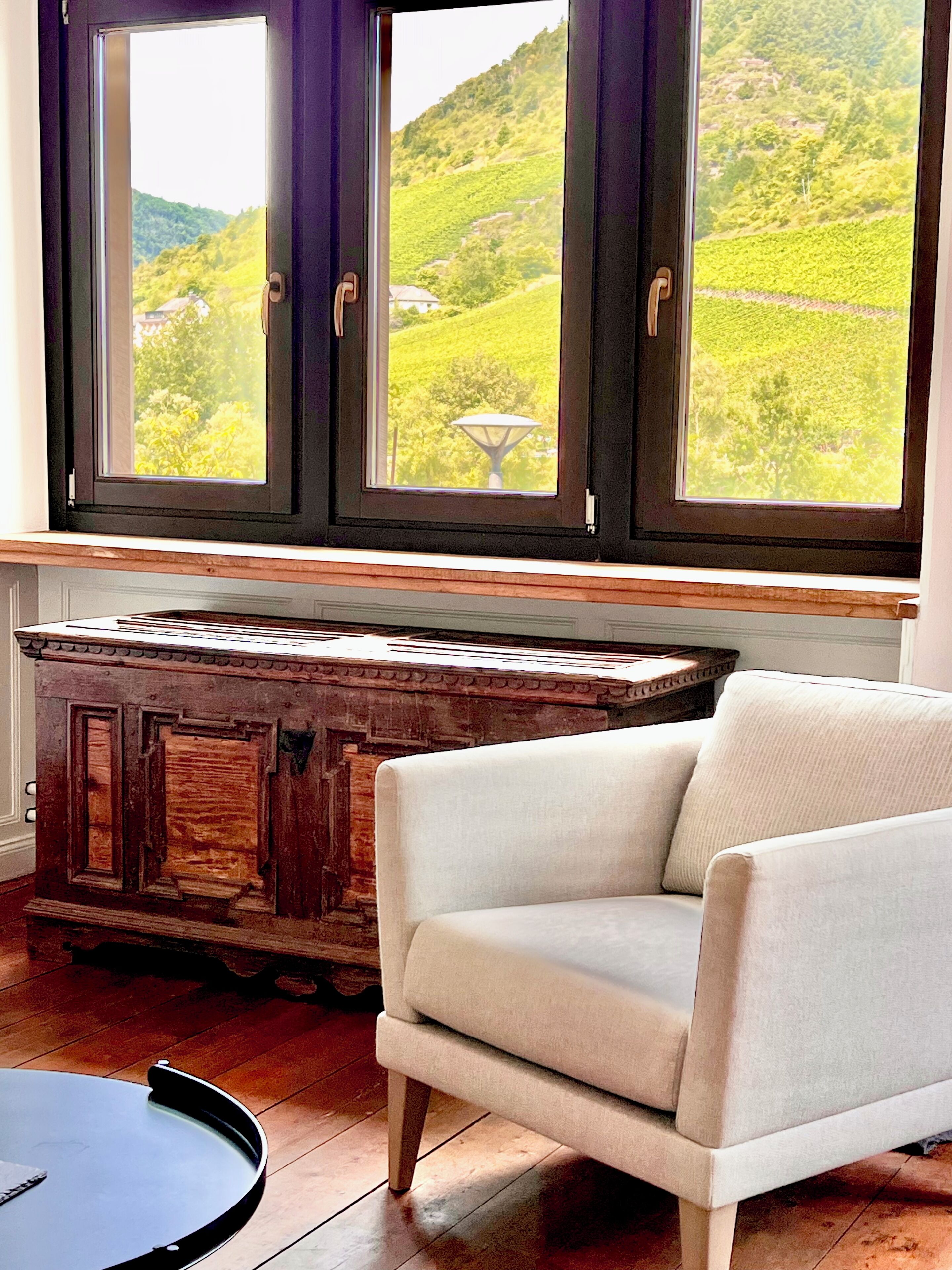 A room with a view of a green landscape through large windows, a wooden cabinet, and a white armchair.