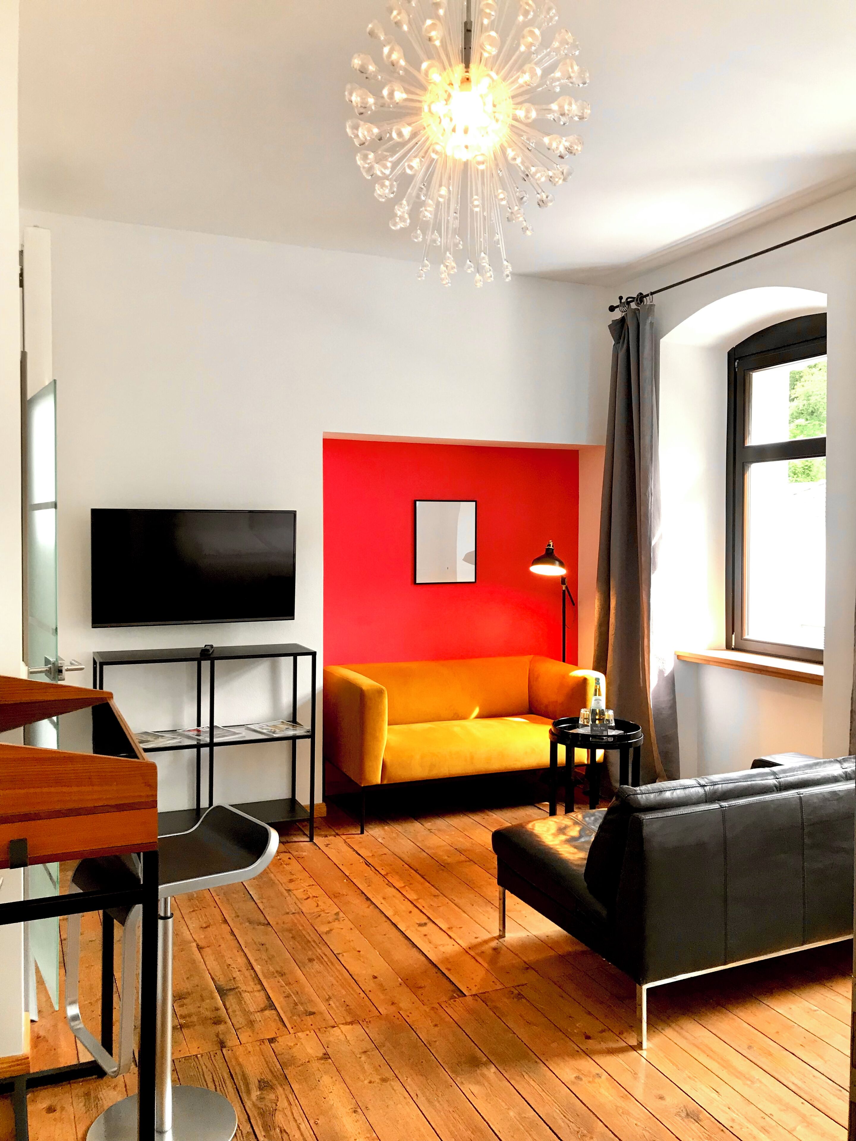 A living room with a TV, a red wall, a wooden floor, a black sofa, and a modern chandelier.