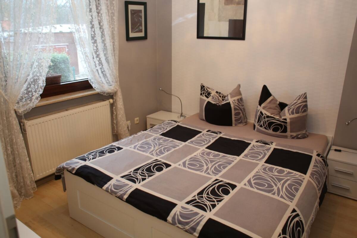 Une chambre à coucher avec un lit, un radiateur, une fenêtre avec des rideaux en dentelle et une table de chevet.