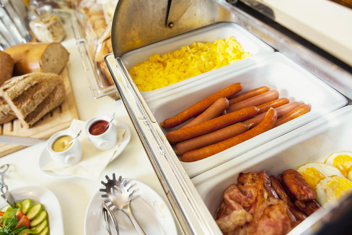A buffet with various food items including sausages, scrambled eggs, and sliced bread.