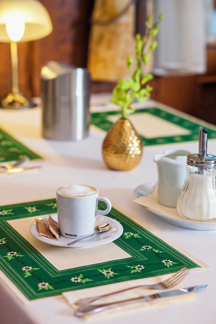 A table set with a cup of coffee, a sugar container, and a pitcher, with a vase of green leaves in the background.