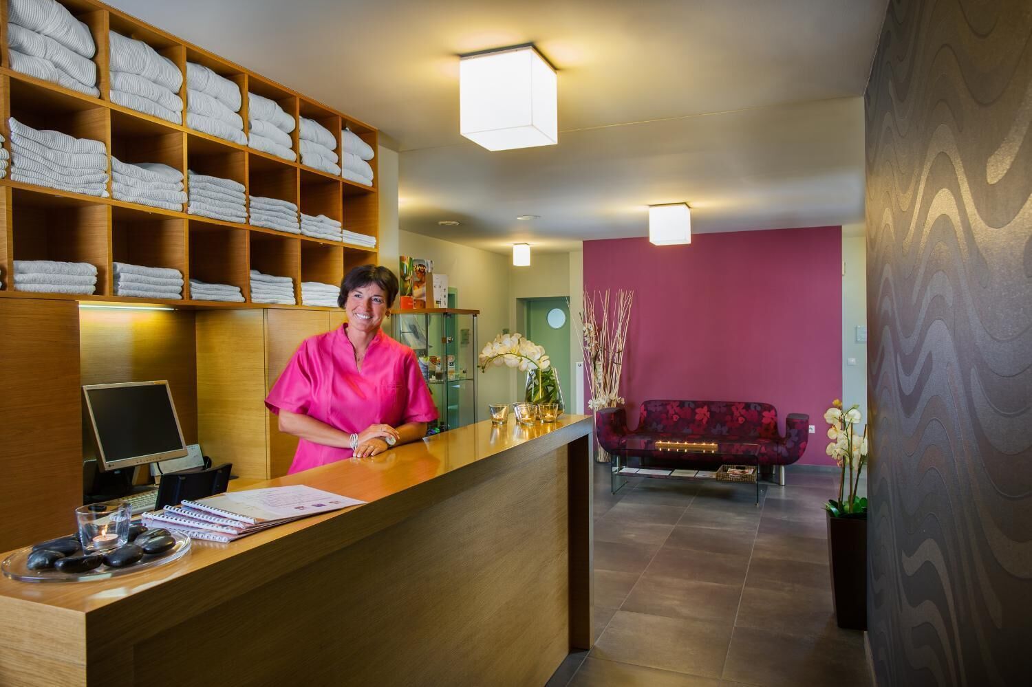 Zona de recepción de un hotel con un mostrador de madera, una computadora y una barra con toallas. Se observa un sofá y un jarrón con flores al fondo.
