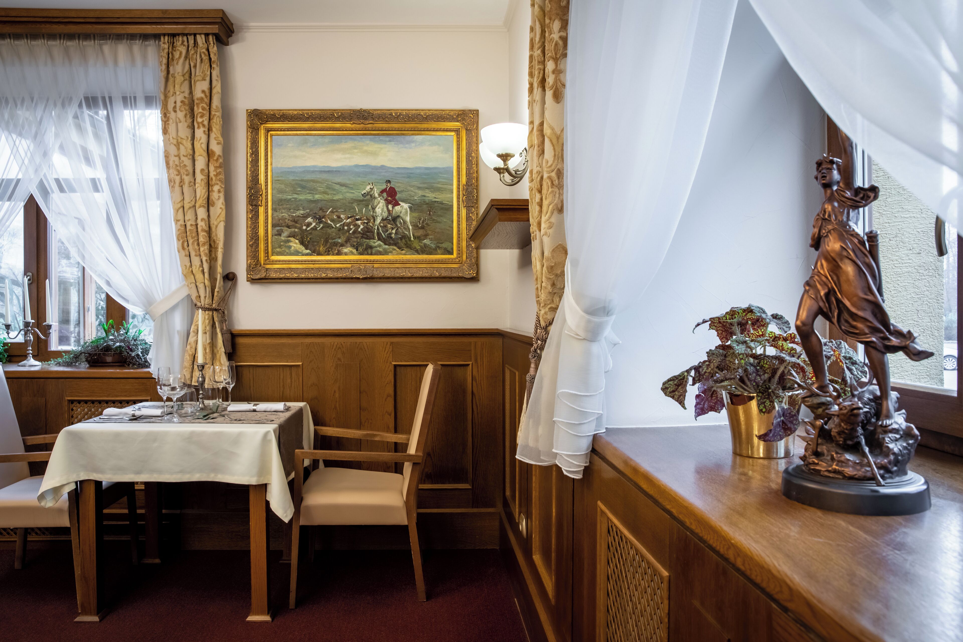A dining area with a table set for two, a framed landscape painting, and a statue on a wooden counter.