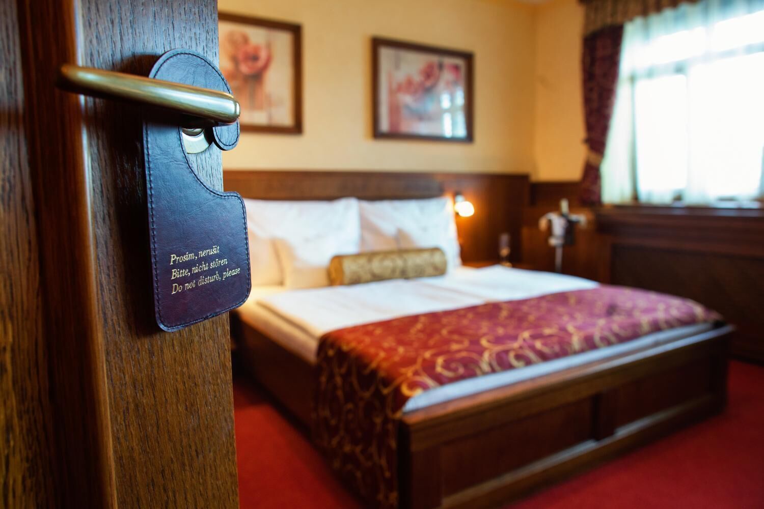 Quarto de hotel com porta de madeira, cama com colcha estampada em vermelho e dourado, e duas fotografias emolduradas na parede.