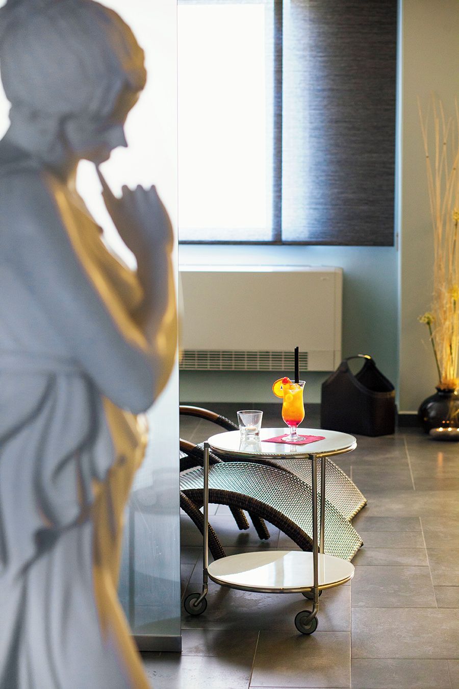 A statue of a figure in a contemplative pose, a small table with a drink, a wicker chair, and a radiator in a room with a window.
