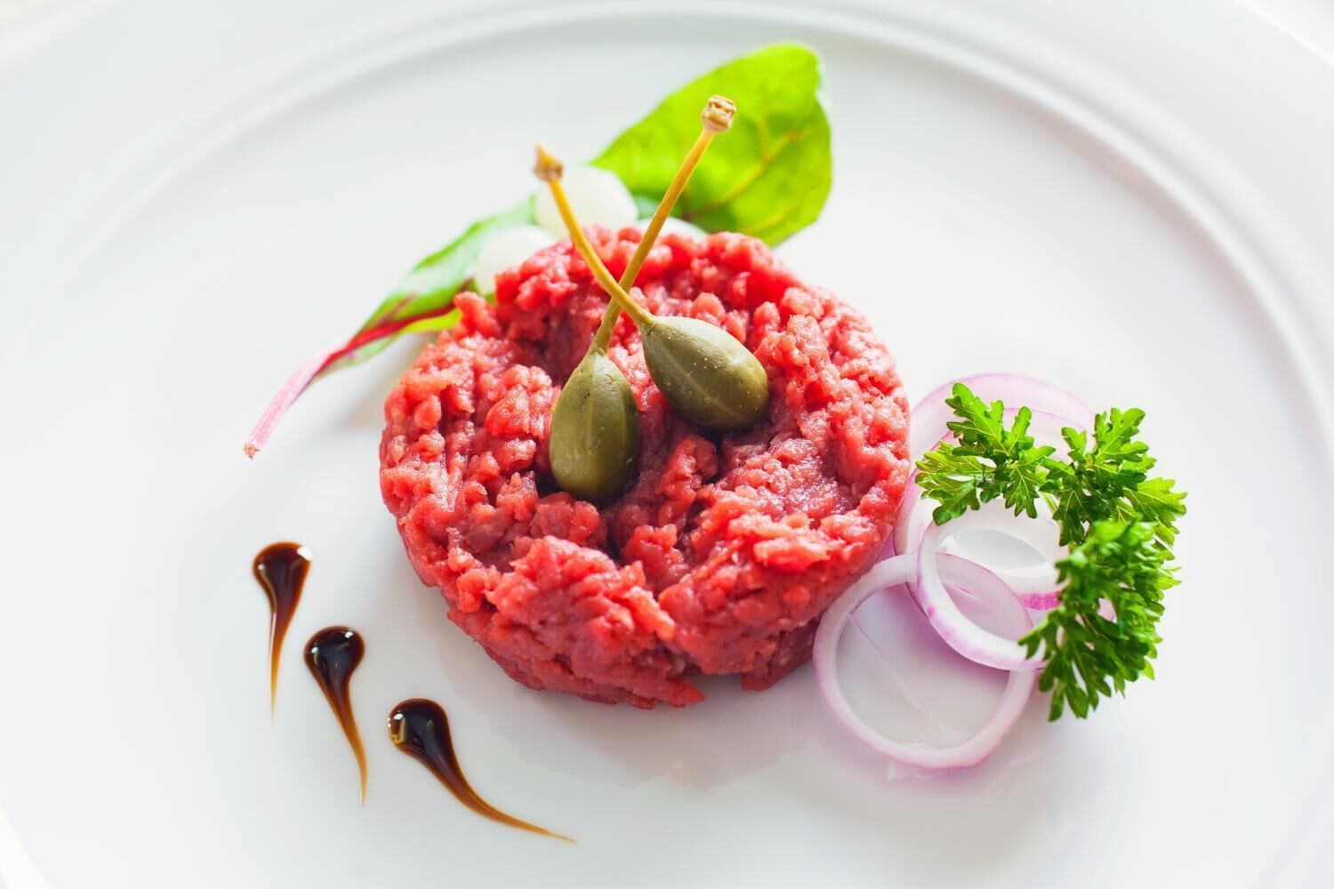 Un plat de tartare de viande crue, agrémenté de câpres et décoré de persil, servi dans une assiette blanche.