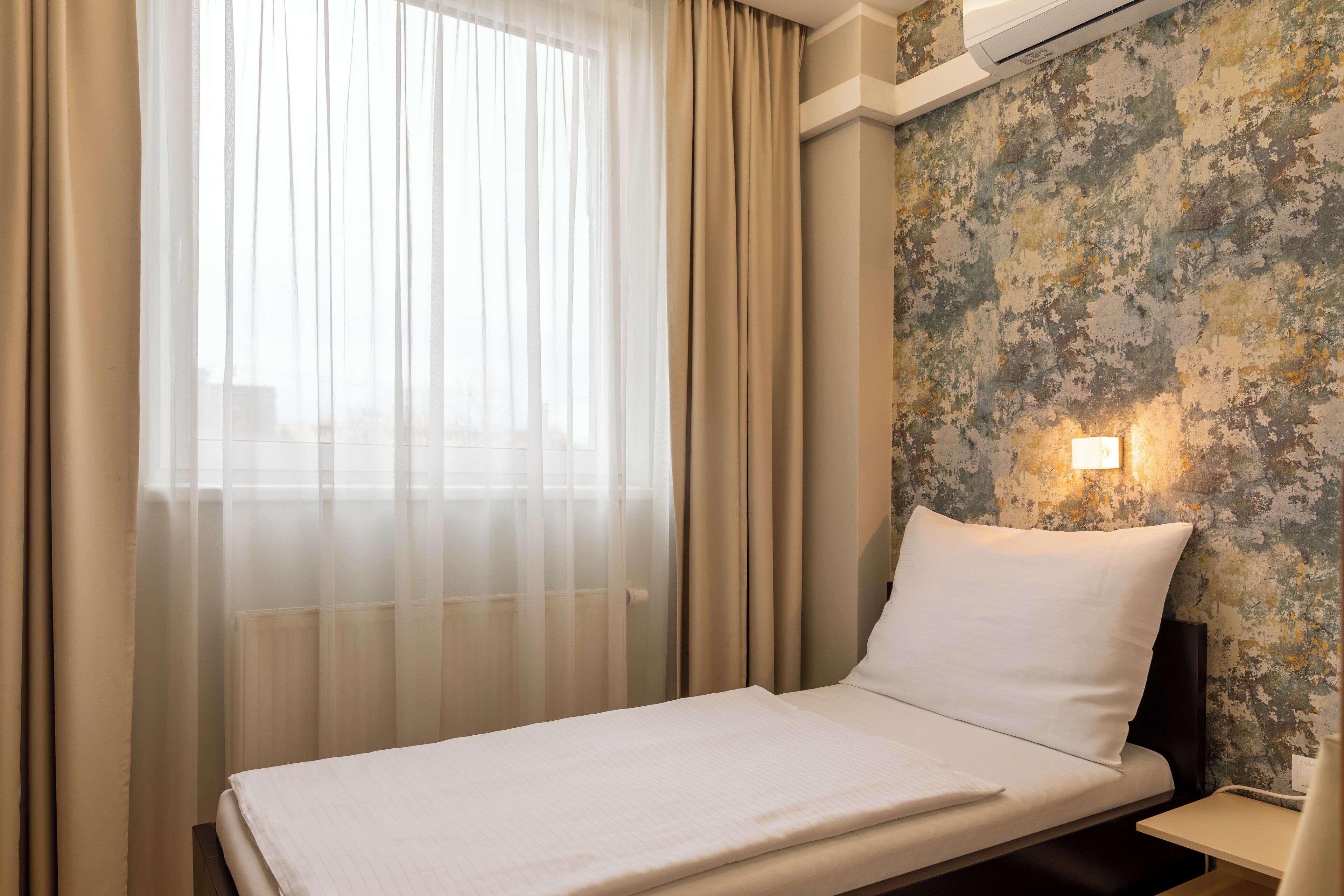 A hotel room with a bed, a window with sheer curtains, a textured wall, and a bedside lamp.