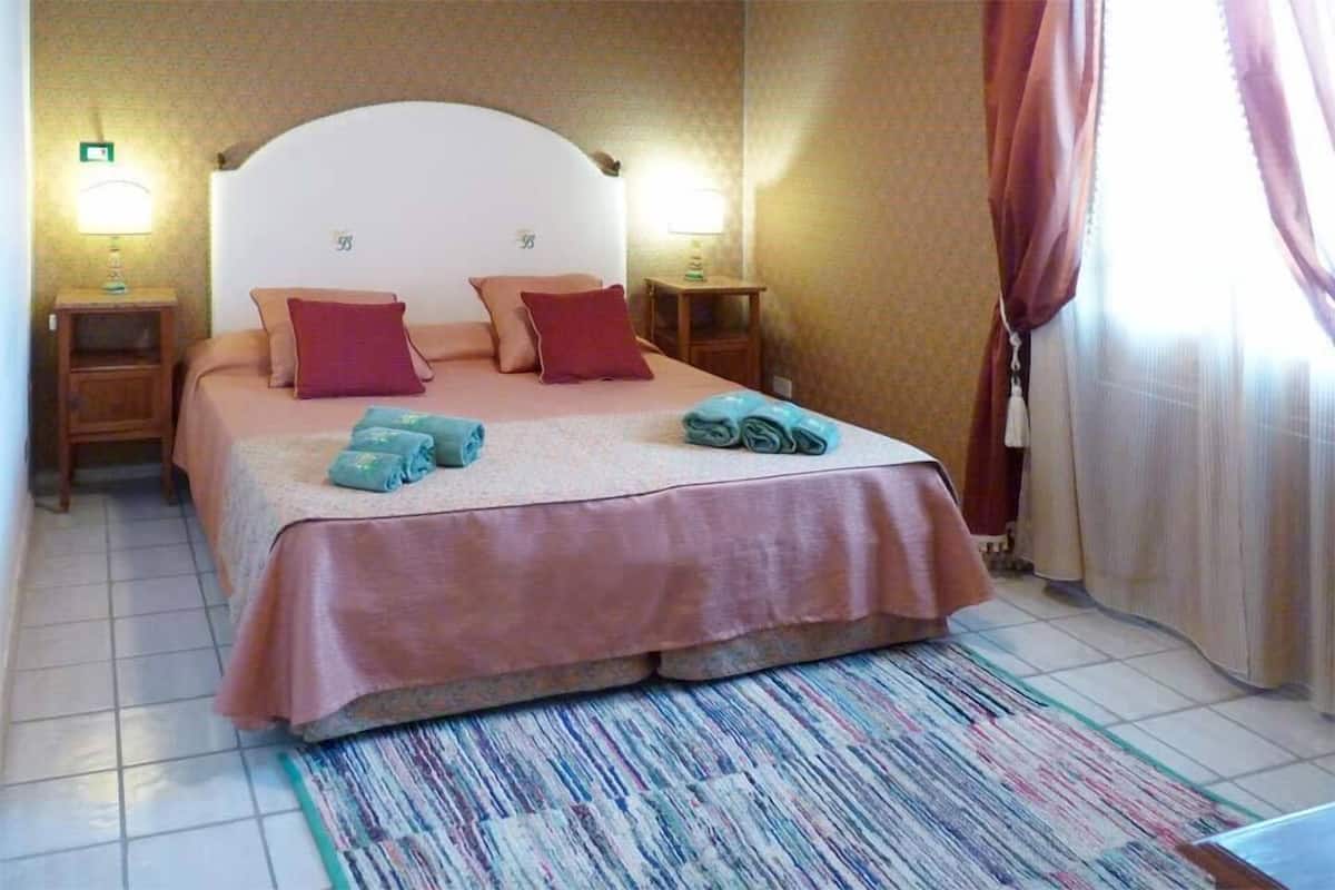 A hotel room with a bed, two bedside tables, a nightstand, a patterned rug, and a window with curtains.