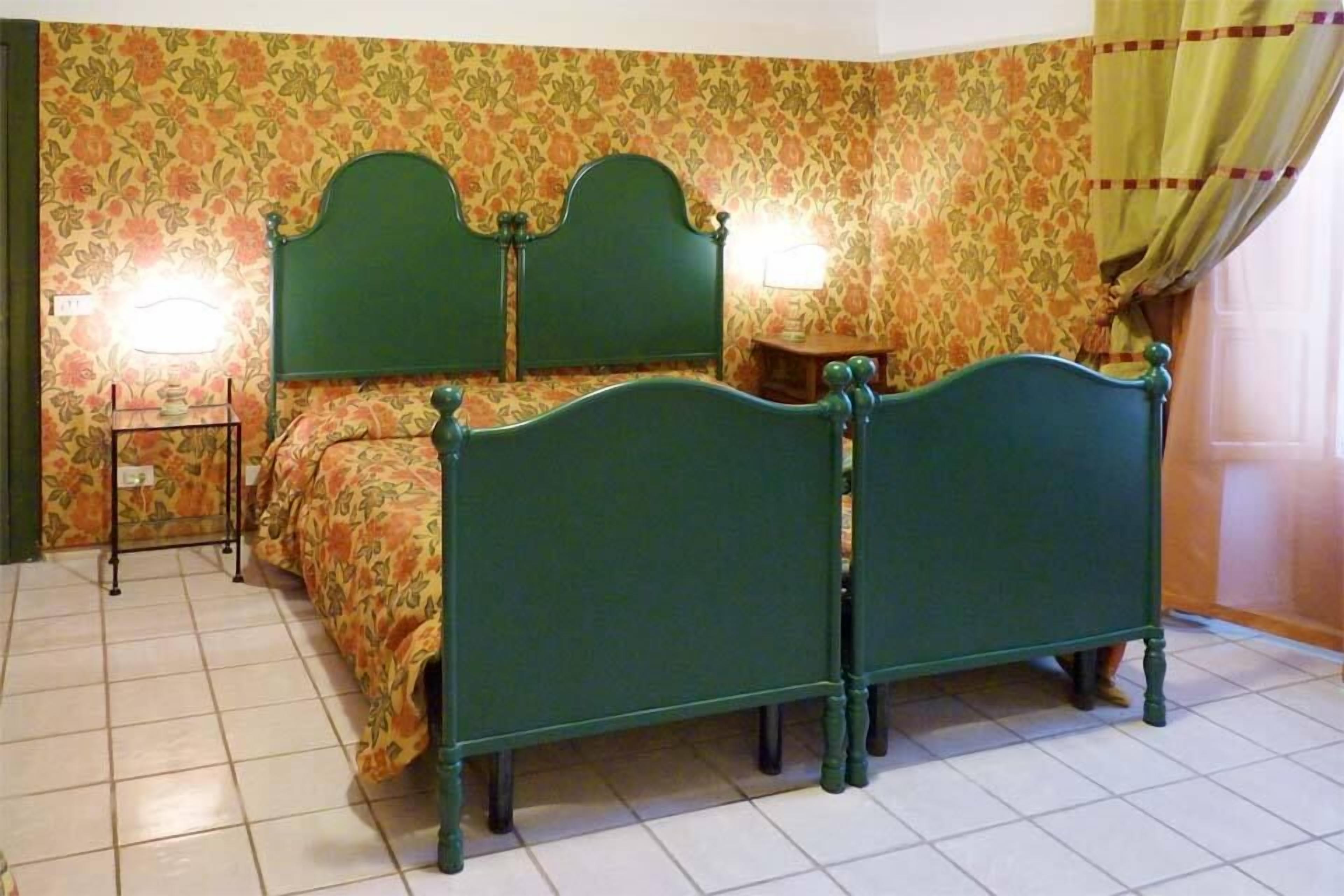 A bedroom with a green upholstered bed, floral wallpaper, and a small bedside table with a lamp.
