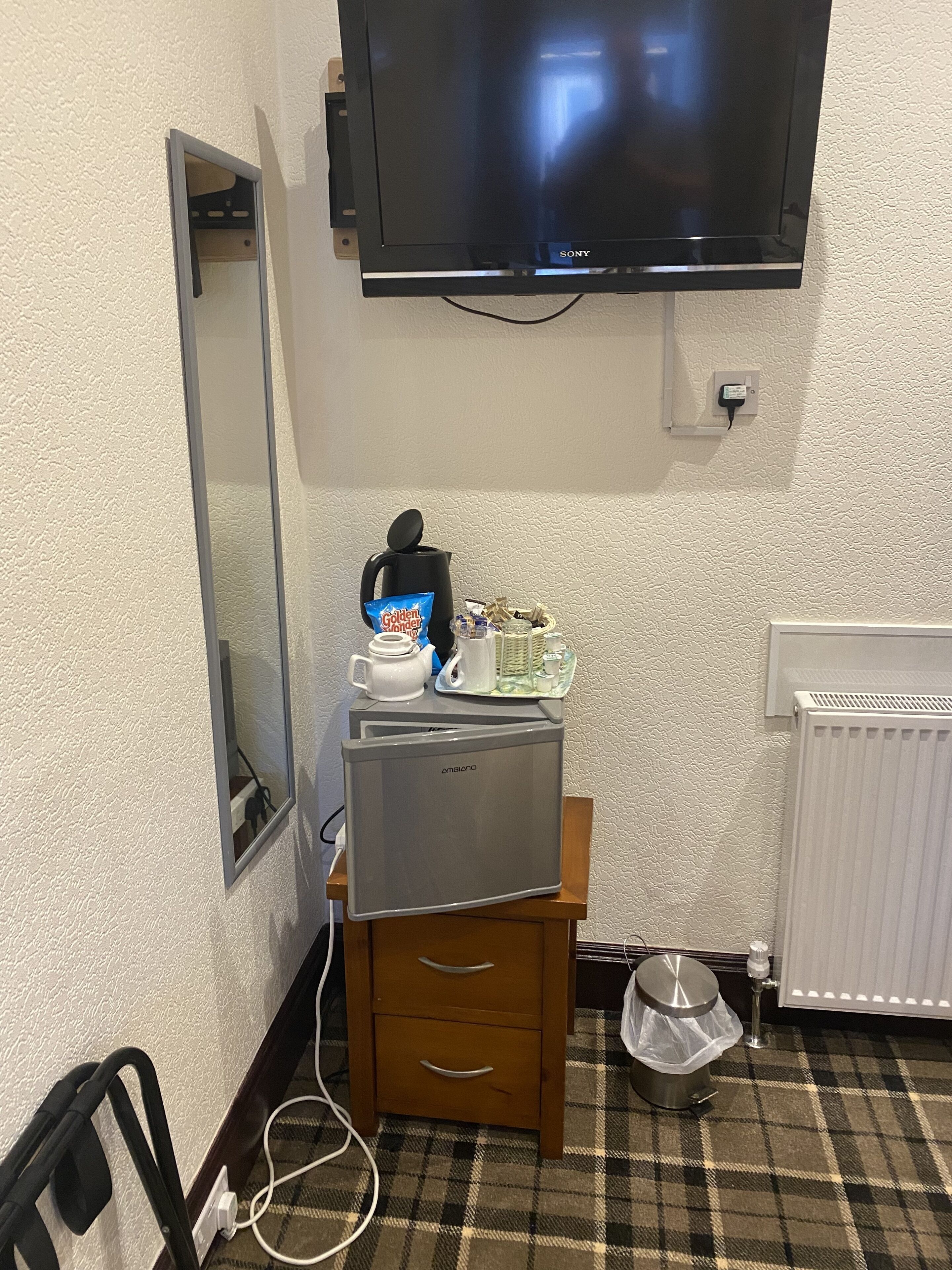 A hotel room with a flat-screen TV mounted on the wall, a minibar with a coffee maker, tea pot, and snacks, a wooden cabinet with drawers, and a radiator on the floor.