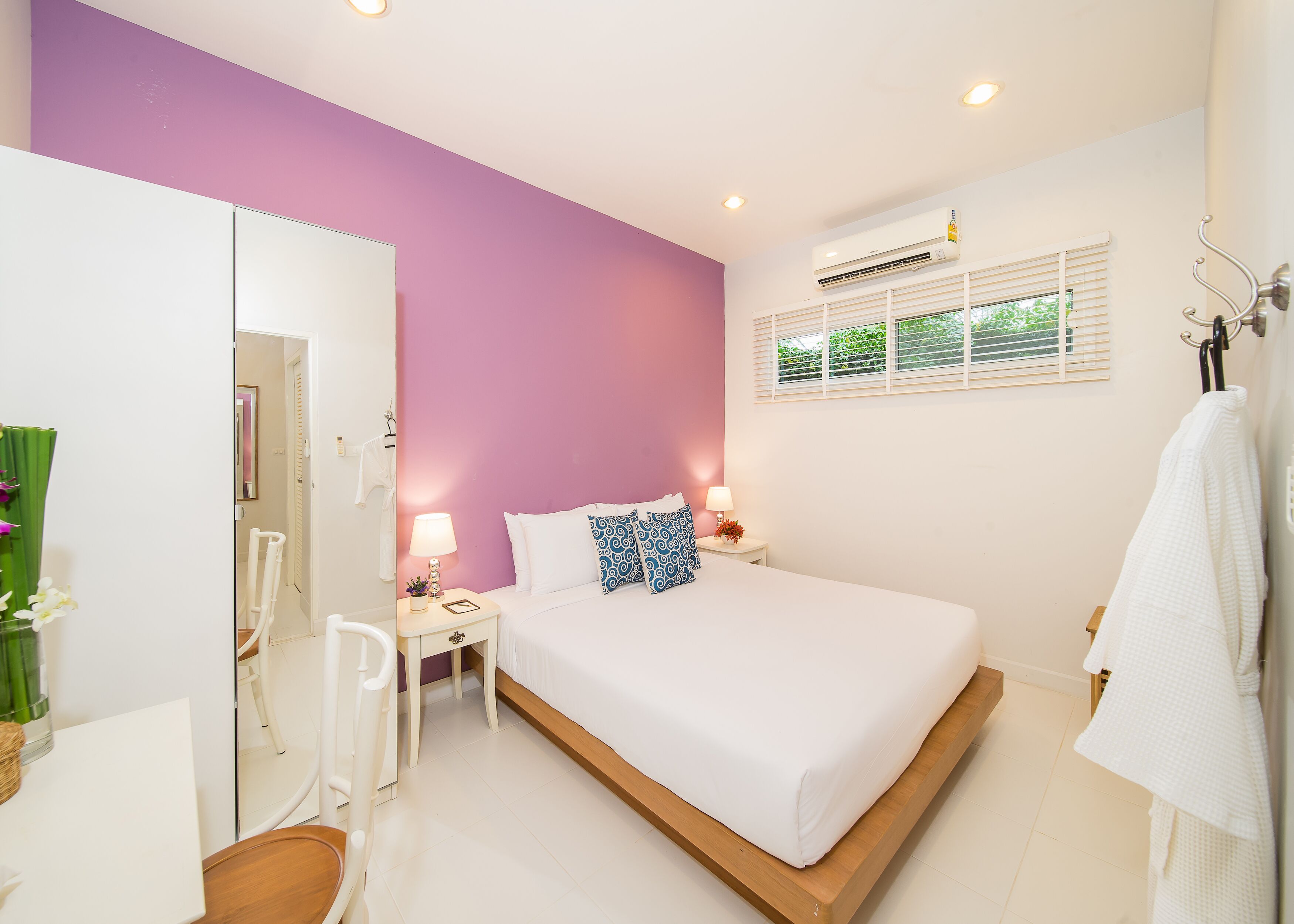 A modern bedroom with a large bed, a bedside table with a lamp, a mirror, and a window with blinds.